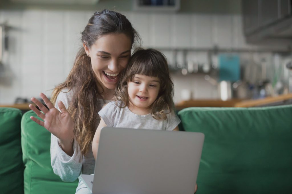 A woman holding a girl on her lap and waving to someone else on a laptop